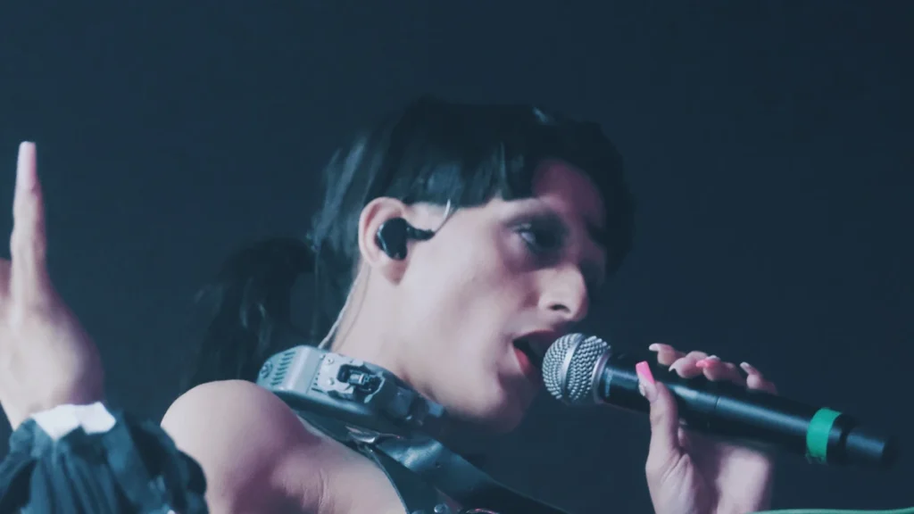 Arca singing into a microphone under blue stage light