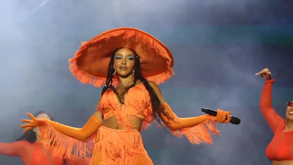 Tinashe performing on stage in an orange fringed outfit and wide-brimmed hat with dancers